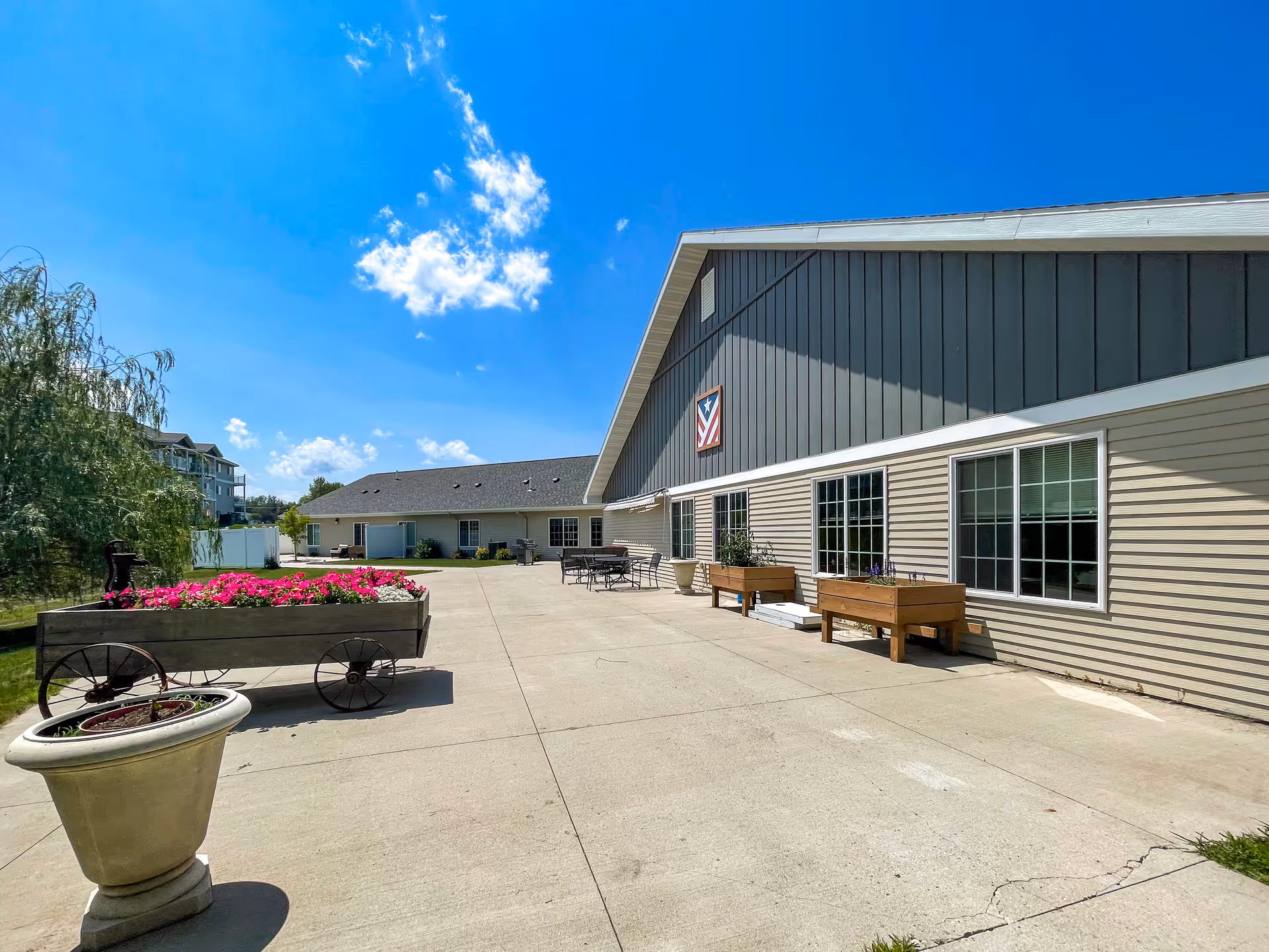 Outdoor patio area at The Legacy Assisted Living Morris, MN, featuring a concrete walkway with flower planters, outdoor seating, and a building with beige siding and a gray upper section under a bright blue sky with some clouds.