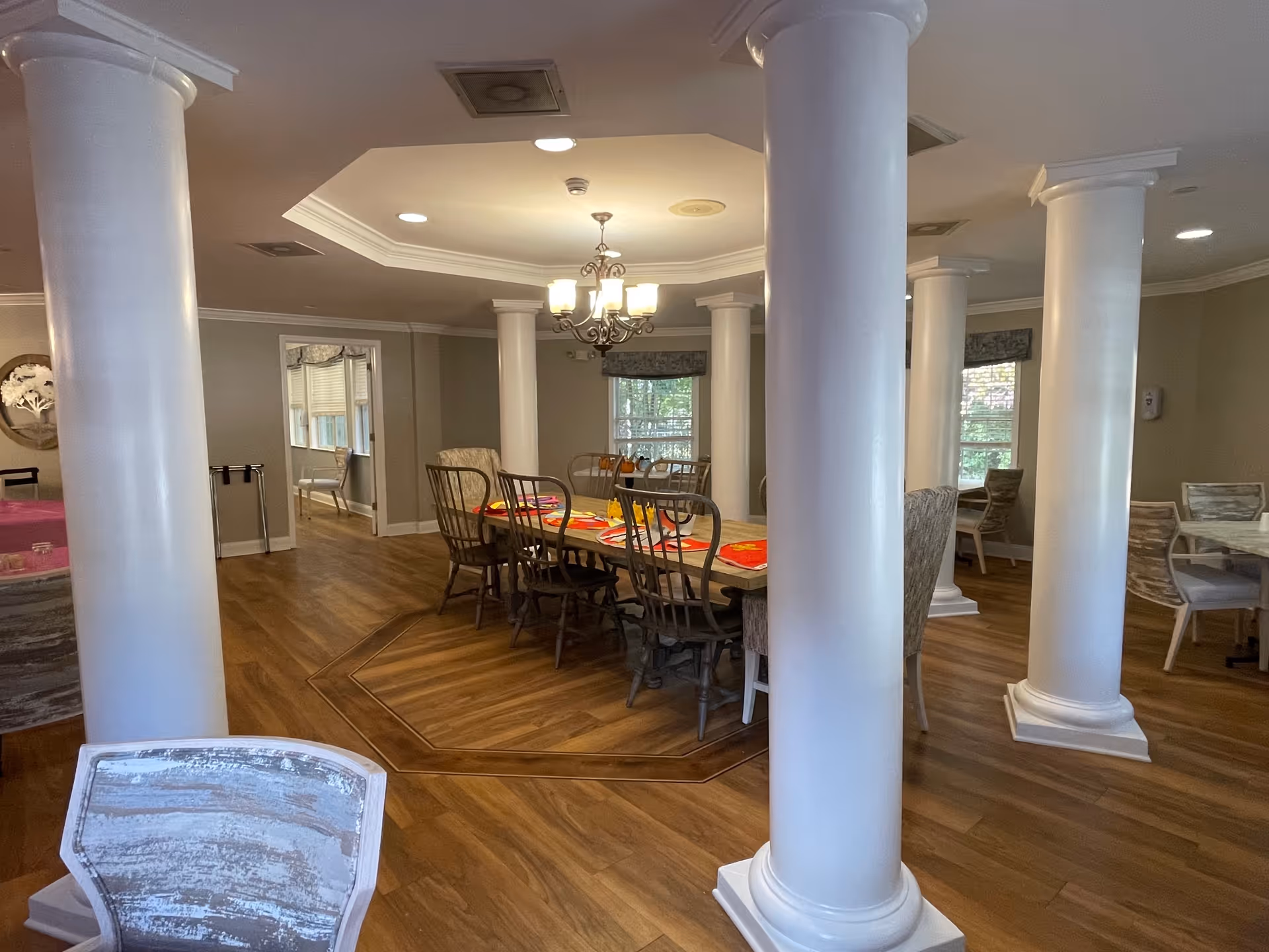 Interior view of a senior living facility dining room with wooden floors, white columns, multiple dining tables with chairs, and a chandelier hanging from the ceiling. The room has windows with valances and neutral-colored walls.