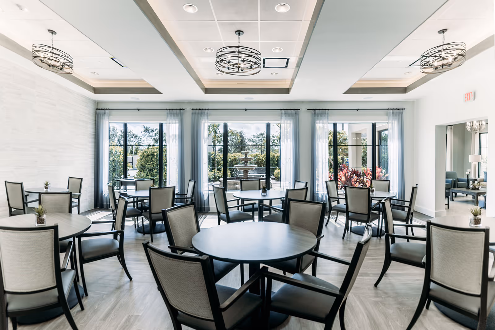 Bright and modern dining room with multiple round tables and cushioned chairs arranged neatly. Large windows with sheer curtains allow natural light to fill the room, and a view of a garden with a fountain is visible outside. Contemporary ceiling lights hang from a recessed ceiling.