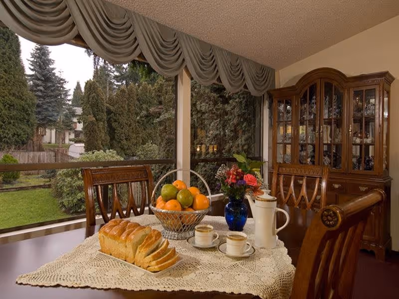 A dining table set with a lace tablecloth, a basket of assorted fruits, a loaf of sliced bread, two cups of coffee with saucers, a white coffee pot, and a blue vase with flowers. The table is surrounded by wooden chairs and is positioned near large windows showing a green garden outside. A wooden china cabinet filled with glassware is visible in the background.