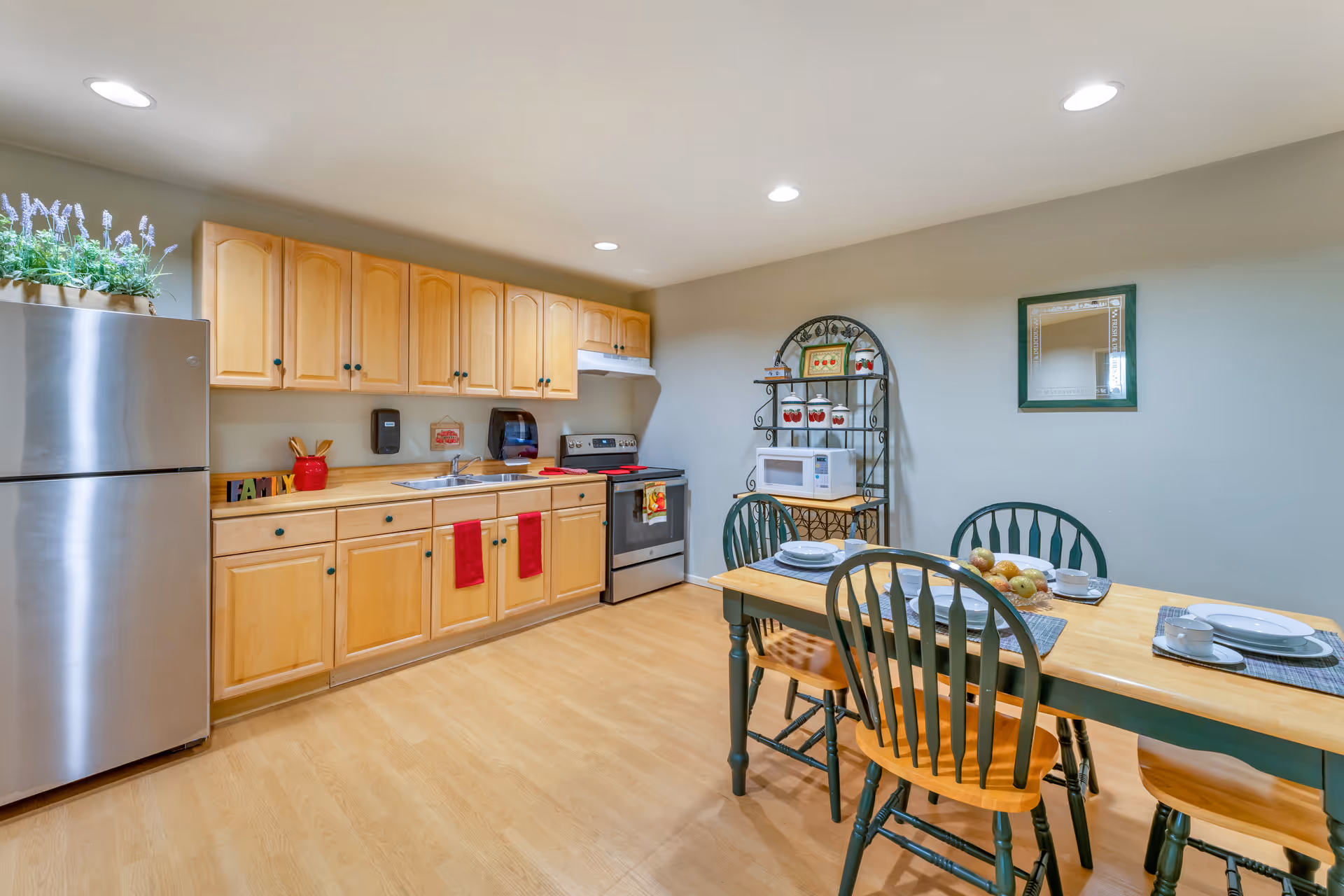 A kitchen and dining area with light wood cabinets, a stainless steel refrigerator, a stove, and a microwave on a decorative metal rack. The dining table is set with plates, cups, and a centerpiece of fruit, surrounded by four green wooden chairs. The room has recessed lighting and light wood flooring.
