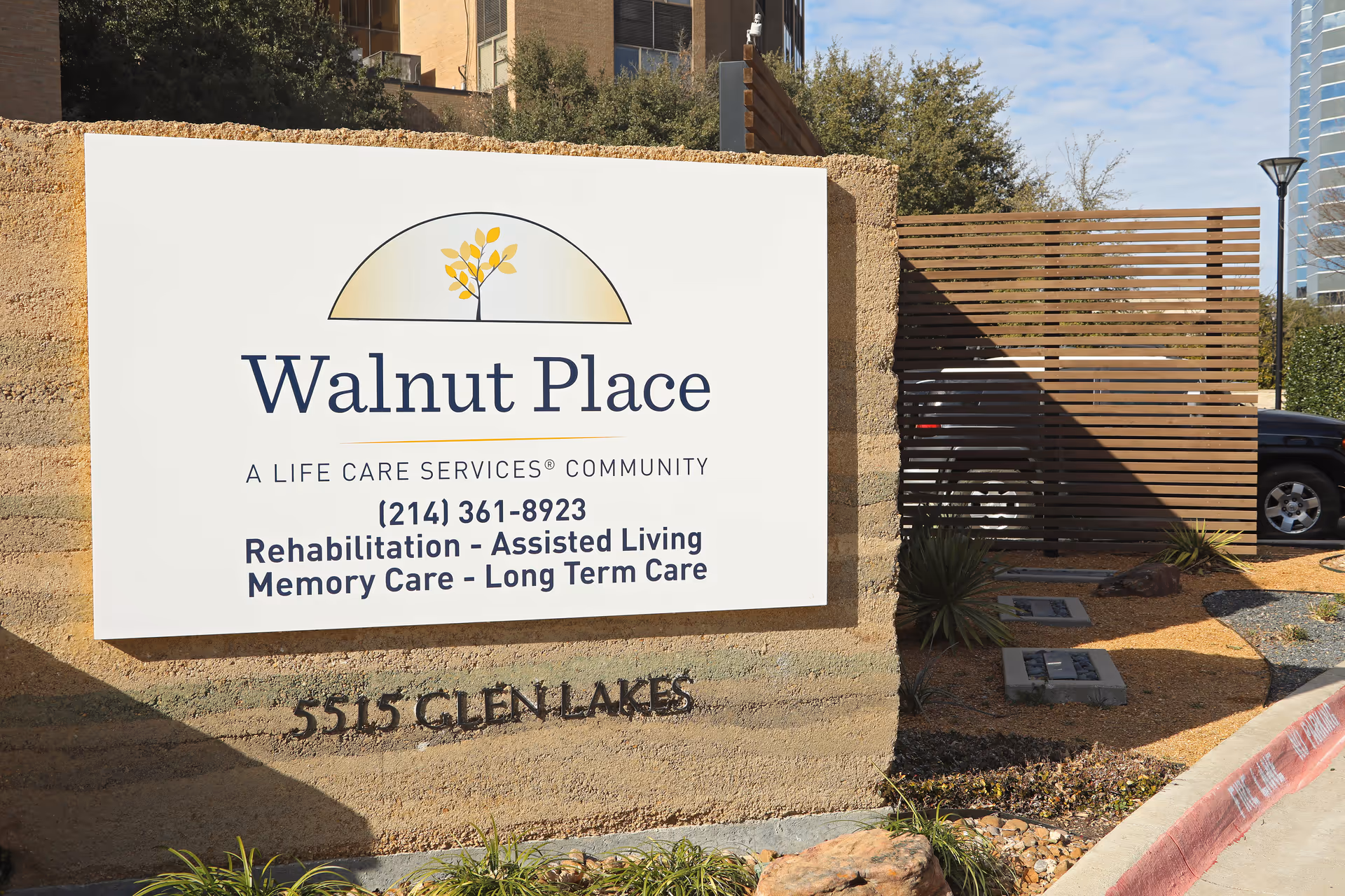 Outdoor sign for Walnut Place, a life care services community offering rehabilitation, assisted living, memory care, and long term care, located at 5515 Glen Lakes. The sign is mounted on a textured stone wall with some plants and a wooden fence in the background.