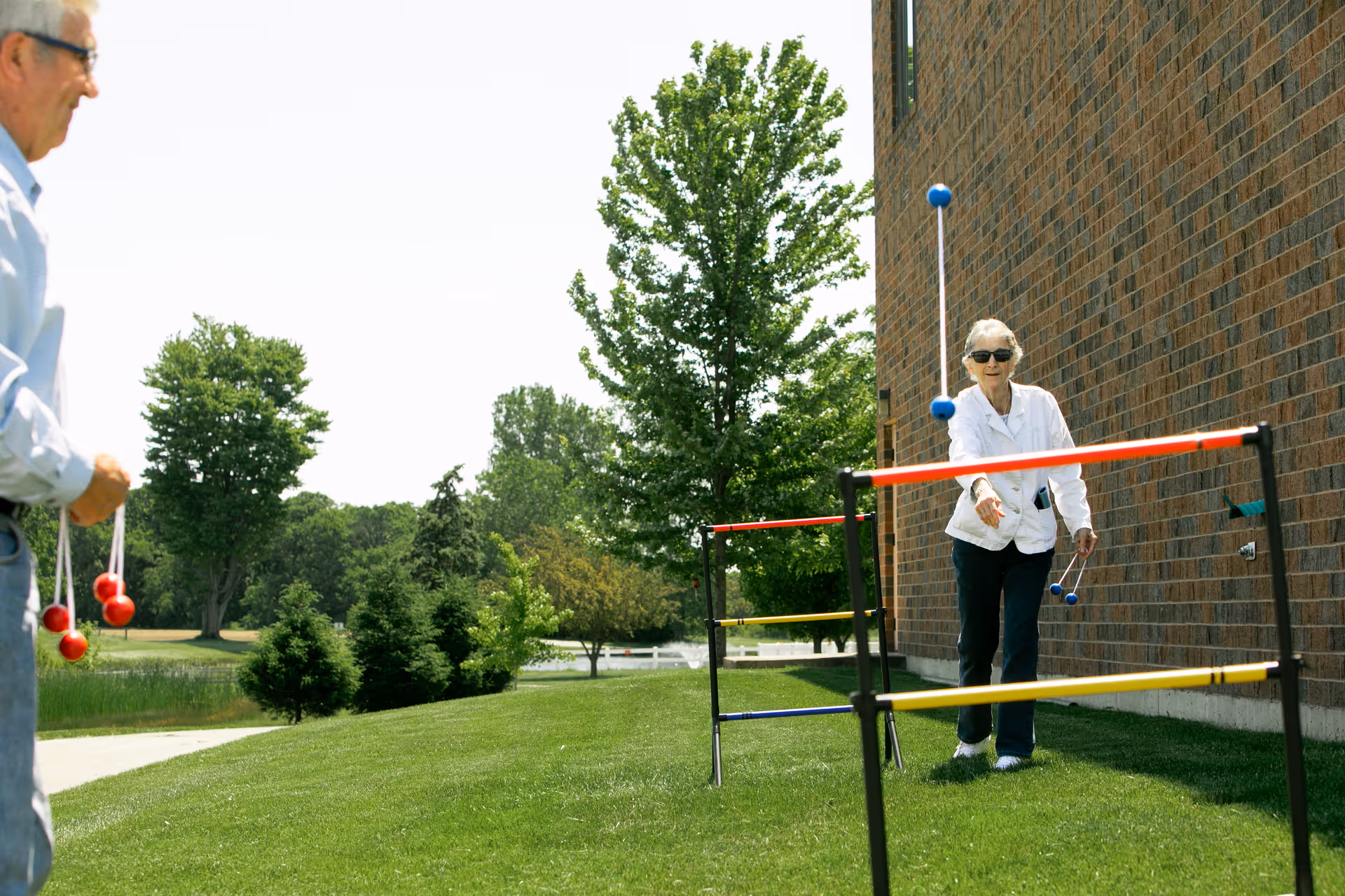 Two seniors playing a ladder toss game on a grassy lawn beside a brick building with trees in the background.