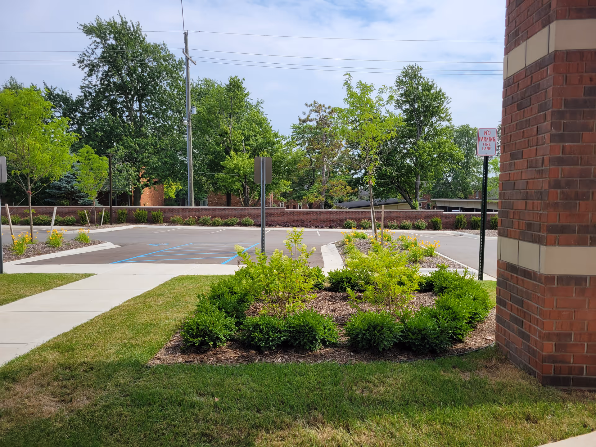 View of a landscaped area with green shrubs and small trees next to a sidewalk and a parking lot. There is a brick building partially visible on the right side and a 'No Parking Fire Lane' sign near the building. Trees and a brick wall are visible in the background under a partly cloudy sky.