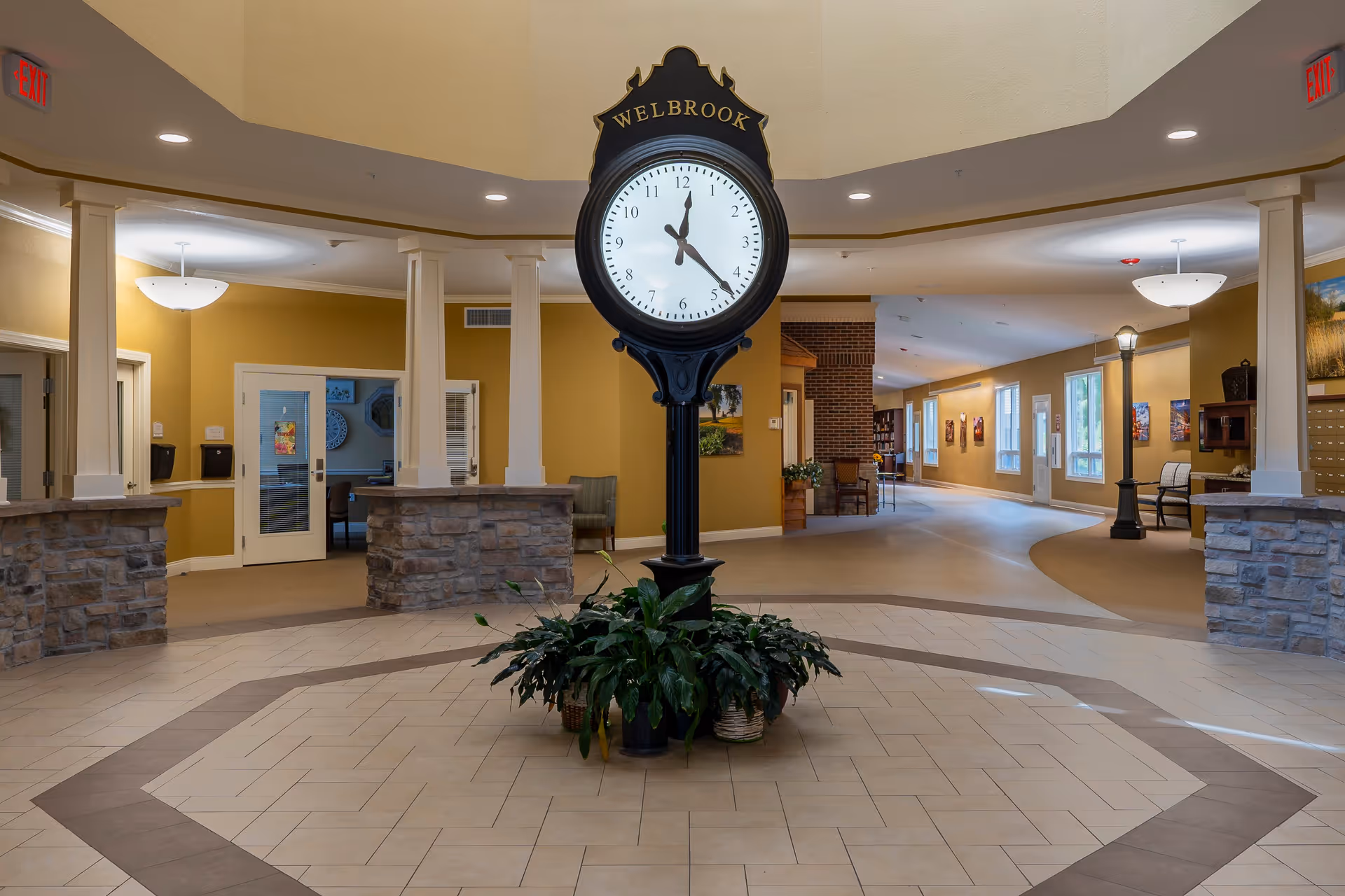 Interior view of a senior living facility lobby area with a large black standing clock labeled 'WELBROOK' in the center surrounded by green plants. The space features beige tiled flooring with a decorative pattern, stone pillars, yellow walls, and multiple doorways leading to other rooms. There are ceiling lights and exit signs visible.