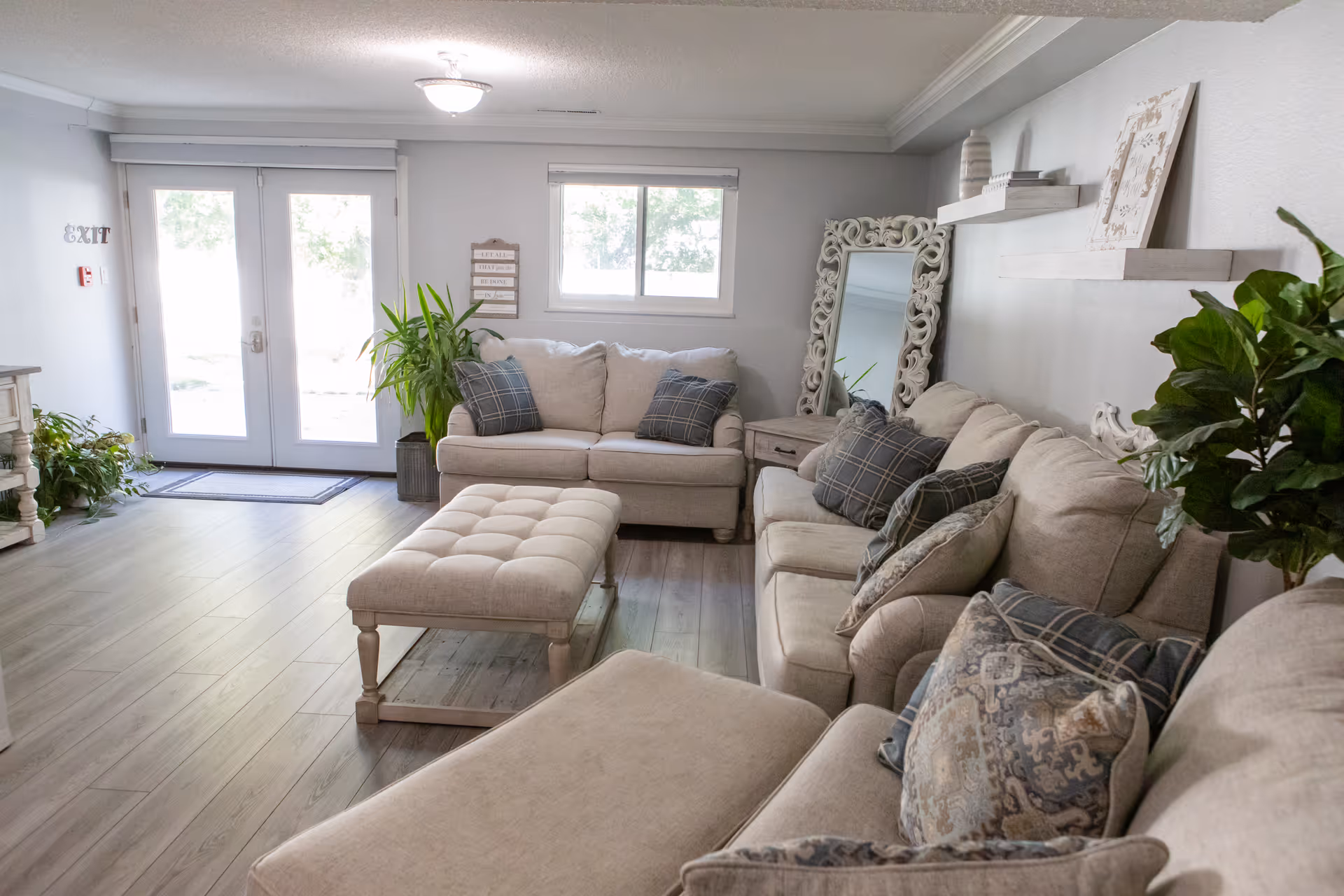 A cozy living room in an assisted living facility featuring beige sofas with patterned cushions, a tufted ottoman, a large ornate mirror leaning against the wall, floating shelves with decorative items, and several green plants. There is a window and glass double doors letting in natural light.
