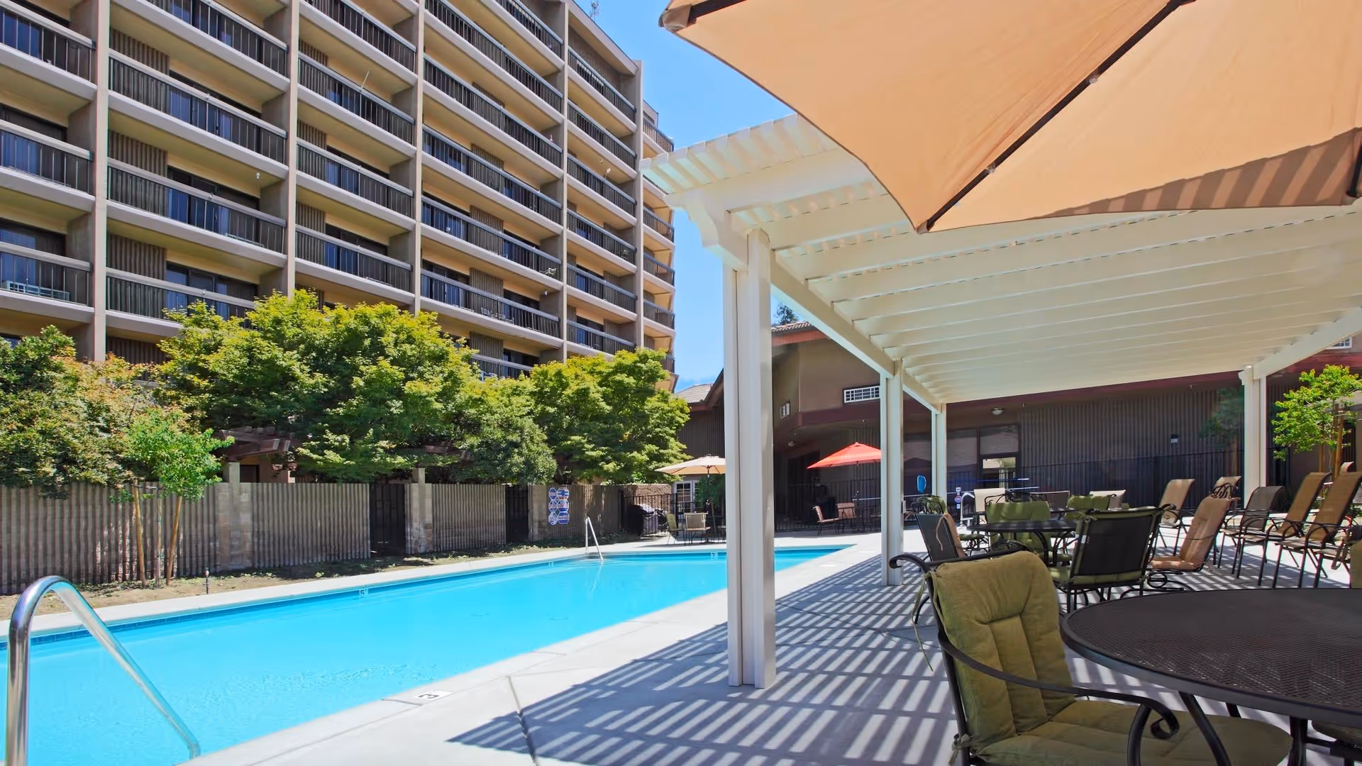Outdoor swimming pool with shaded patio seating and lounge chairs beside a multi-story retirement community building.
