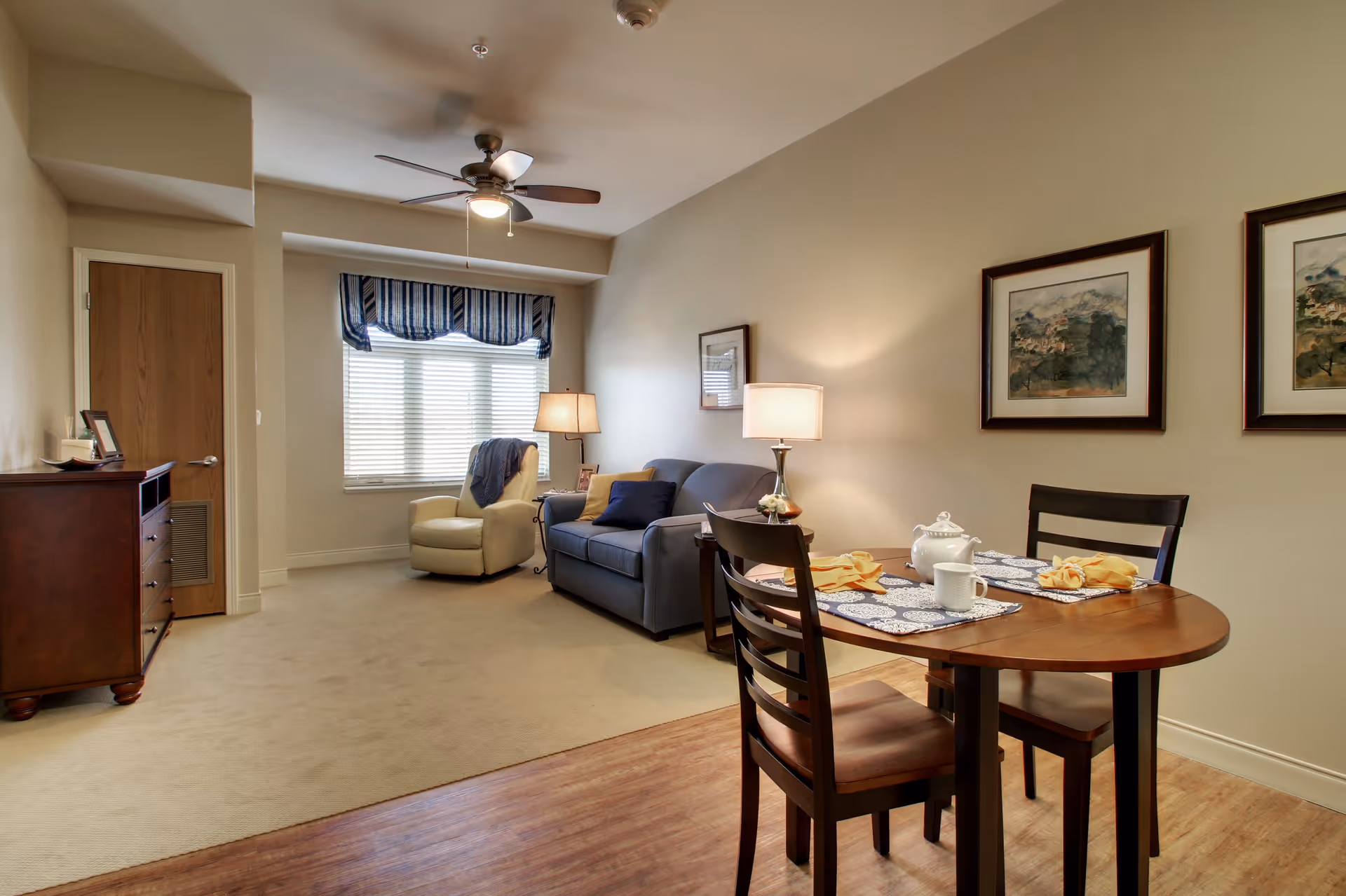 A cozy senior living room with a small dining table set for two, a blue sofa, a beige armchair near a window with blinds and a striped valance, two framed paintings on the wall, a ceiling fan with light, and a wooden dresser near a closed door.
