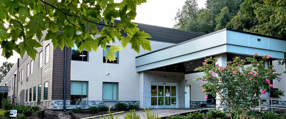 Exterior view of a modern two-story healthcare facility building with large windows, a covered entrance, and landscaped greenery including trees and flowering bushes in the foreground.