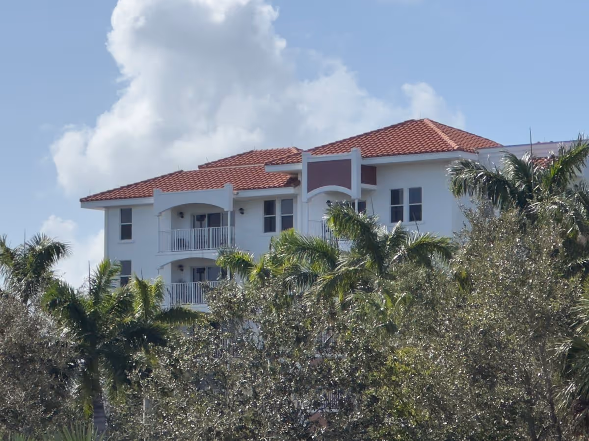 White multi-story building with a red tile roof and balconies partially obscured by palm trees under a blue sky.