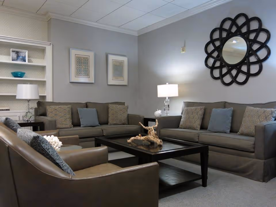 A cozy living room with two gray sofas and two brown armchairs arranged around a dark wooden coffee table with a decorative driftwood centerpiece. The walls are painted light gray and decorated with two framed pictures and a large black decorative mirror. There are two table lamps on side tables providing soft lighting, and a built-in white bookshelf with decorative items in the background.