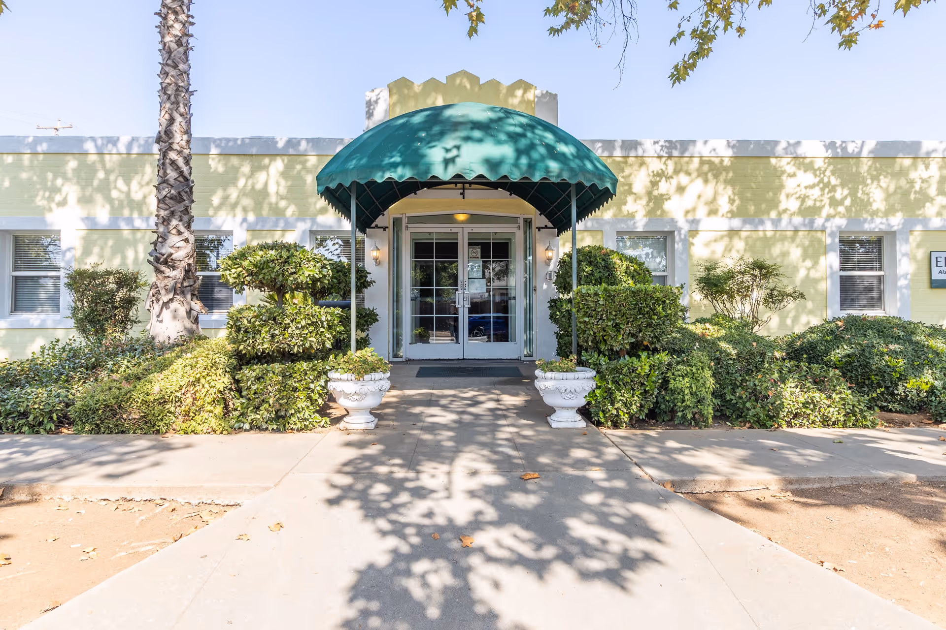 Front entrance of a single-story building with a green awning above double glass doors, surrounded by neatly trimmed bushes and a palm tree, under a clear blue sky.