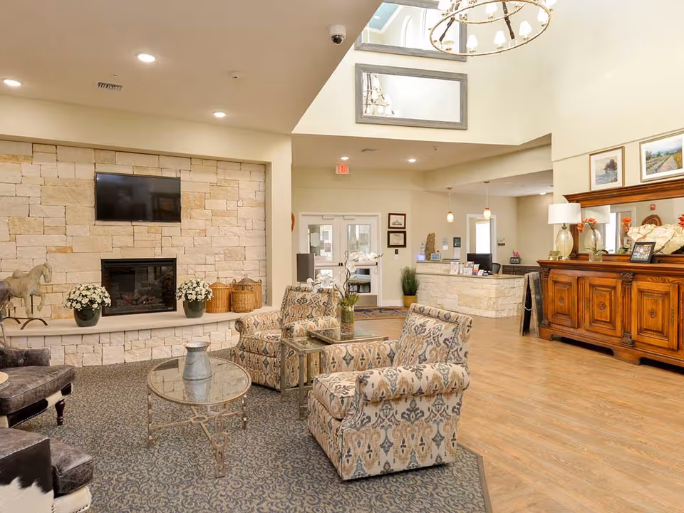 A cozy senior living facility common area with patterned armchairs and a glass coffee table on a carpeted floor. There is a stone fireplace with a mounted TV above it, decorative plants, and baskets on the hearth. The room features a wooden sideboard with lamps and framed pictures, a reception desk in the background, and a high ceiling with a chandelier and skylight.