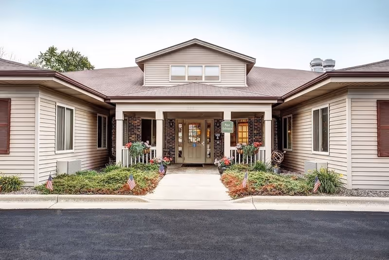 Front exterior view of a single-story building with beige siding and a covered entrance. The entrance has a sign that reads 'Main Entrance' and is flanked by flower pots and small American flags along the walkway. The roof has a small dormer window, and there are shrubs and landscaping on either side of the path leading to the door.