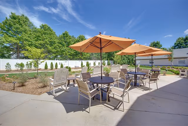 Outdoor patio area with several round tables, each shaded by large orange umbrellas. The tables are surrounded by beige chairs. The patio is adjacent to a garden area with small trees and shrubs, enclosed by a white fence. The sky is clear and blue with some wispy clouds.