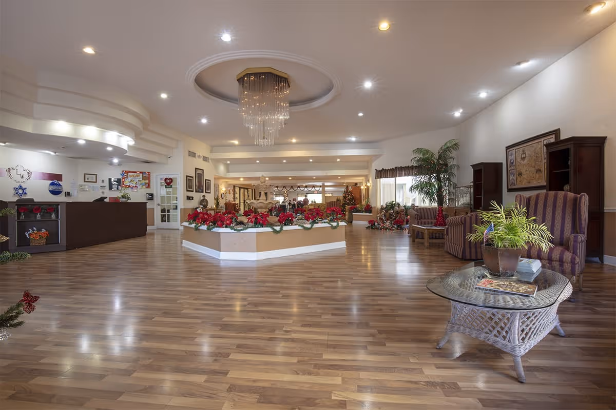 Spacious senior living facility lobby with polished wooden floors, a central planter decorated with red poinsettias, a chandelier hanging from a recessed ceiling, a reception desk on the left, and comfortable seating areas with armchairs and a glass-top table on the right. The space is decorated for the holidays with a Christmas tree and festive ornaments.