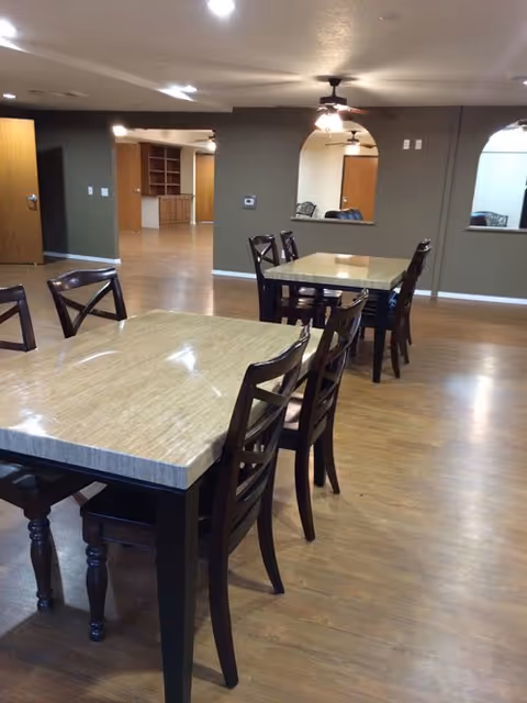 Interior view of a senior living facility dining area with two rectangular tables and several wooden chairs on a wood floor. The room has beige walls with arched openings and ceiling fans with lights.