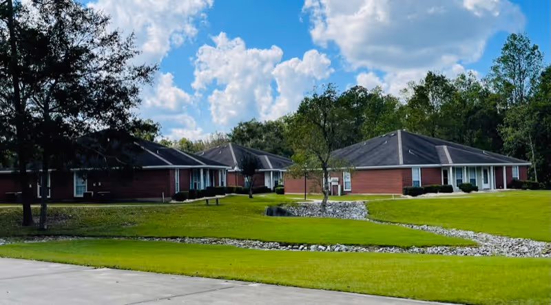 View of a senior living community with single-story brick buildings surrounded by green lawns, trees, and a partly cloudy blue sky.