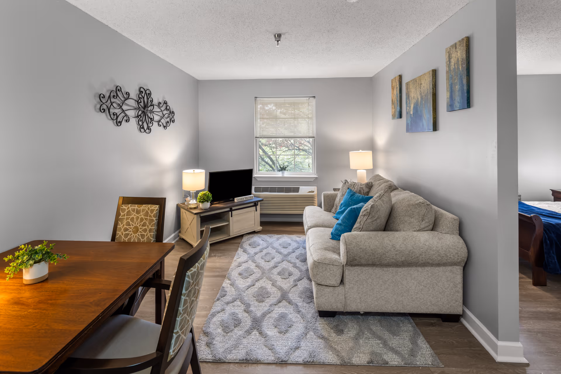 A cozy living room area with a beige sofa adorned with blue pillows, a patterned gray rug, a wooden TV stand with a small flat-screen TV, two table lamps, and a window with blinds. Adjacent to the living room is a wooden dining table with two chairs, and part of a bedroom with a bed covered in blue bedding is visible through an open doorway.