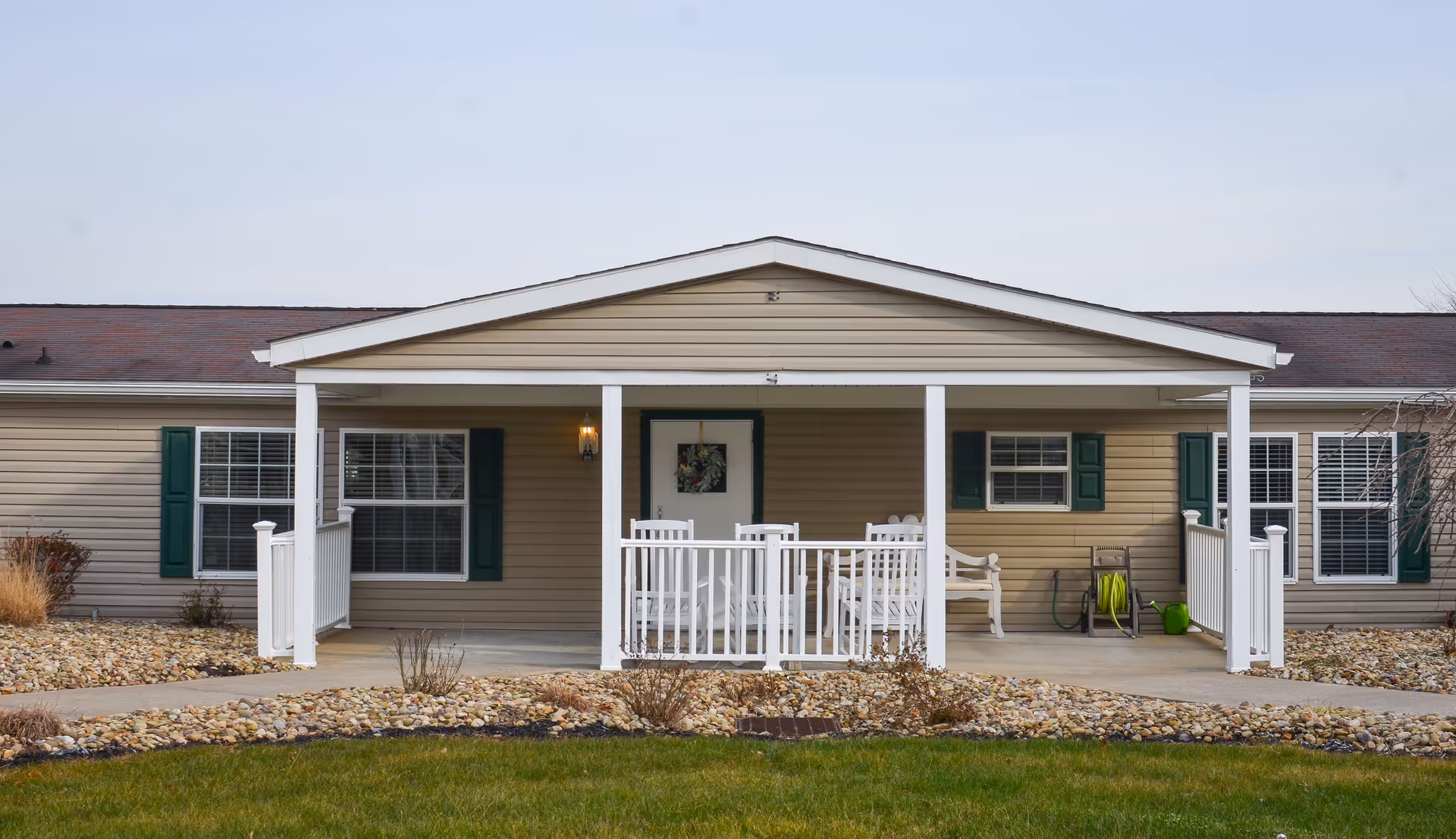 Front exterior view of a single-story senior living facility building with beige siding, green window shutters, and a covered porch with white railings and outdoor seating. The porch has a light fixture and a decorative wreath on the door. The surrounding area includes a rock garden and a grassy lawn.