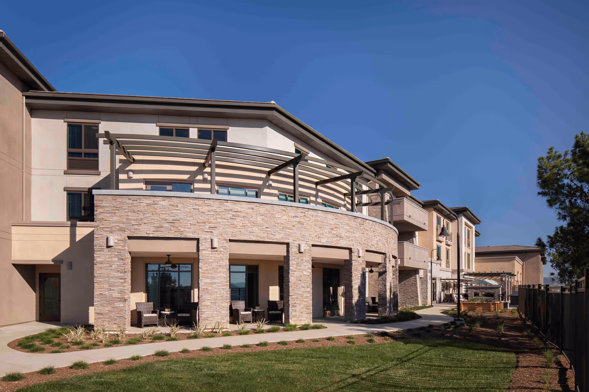 Exterior view of a modern senior living facility building with stone and stucco facade, featuring a covered patio area with seating and a pergola on the upper balcony, surrounded by a landscaped lawn and clear blue sky.
