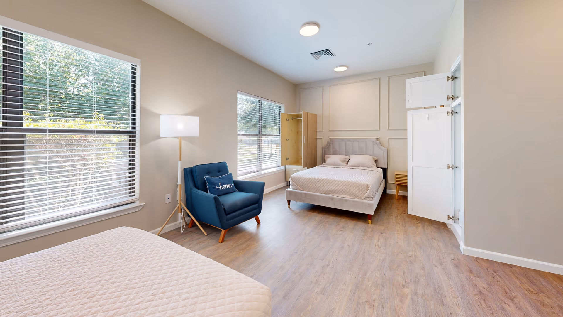 Spacious, well-lit bedroom with two beds, a blue armchair, large windows, and wood-look flooring.
