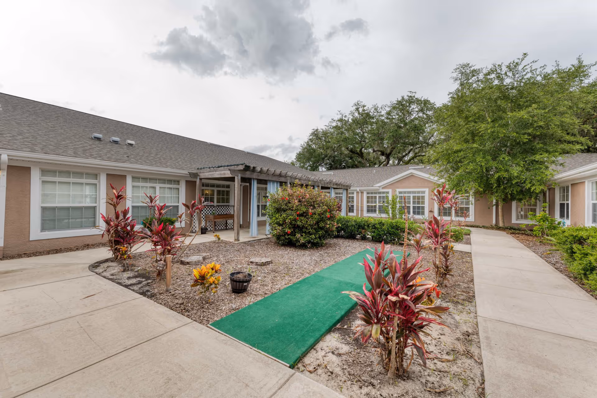 Outdoor courtyard area at Addington Place of Titusville featuring a paved walkway, landscaped garden beds with red and yellow plants, a green carpeted path, and a building with large windows and a covered pergola.