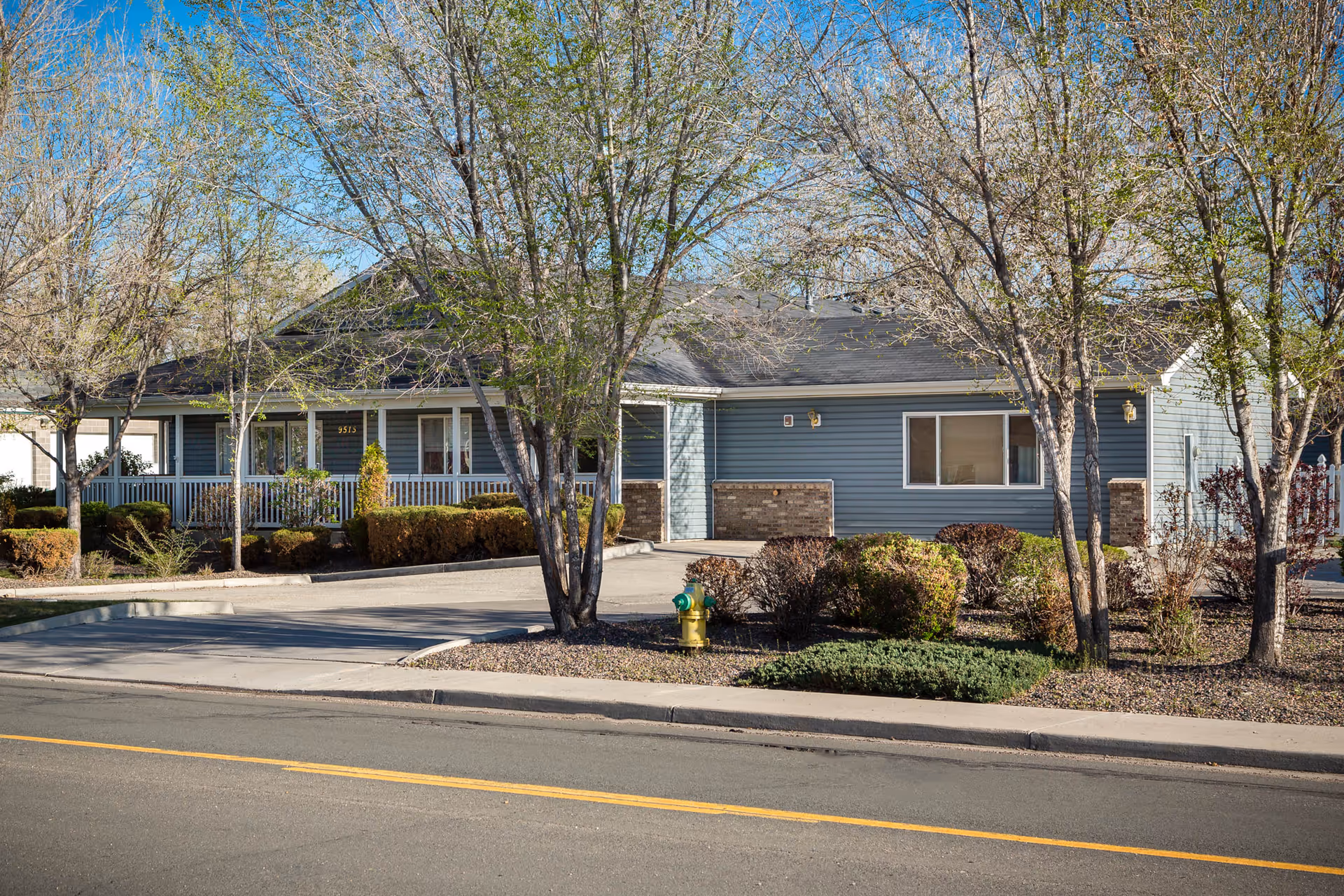 Single-story assisted living building front with a covered porch, trees, landscaping, and a yellow fire hydrant by the road.