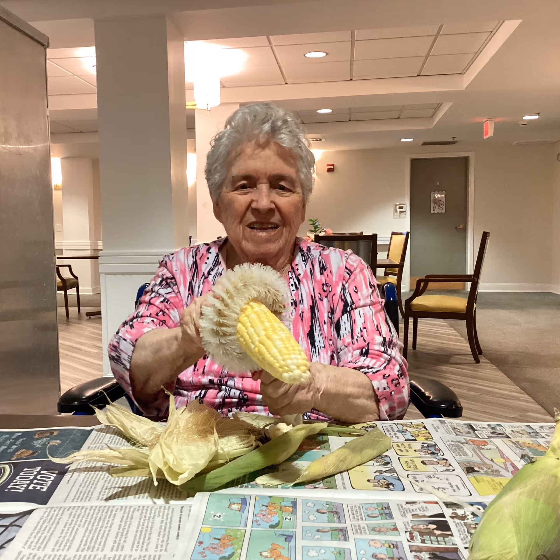 An elderly woman with short gray hair wearing a pink patterned shirt is sitting at a table in a well-lit room. She is smiling and holding an ear of corn with a corn brush, cleaning or husking it. The table is covered with newspaper and several ears of corn with husks are visible. In the background, there are chairs and a door, suggesting a communal indoor space.