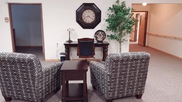 A seating area with two patterned armchairs facing a small wooden table. Behind the table is a wooden desk with a chair, a lamp, a decorative plate, and a large wall clock above it. To the right of the desk is a tall green plant. The area is carpeted and there are doorways and a hallway visible in the background.
