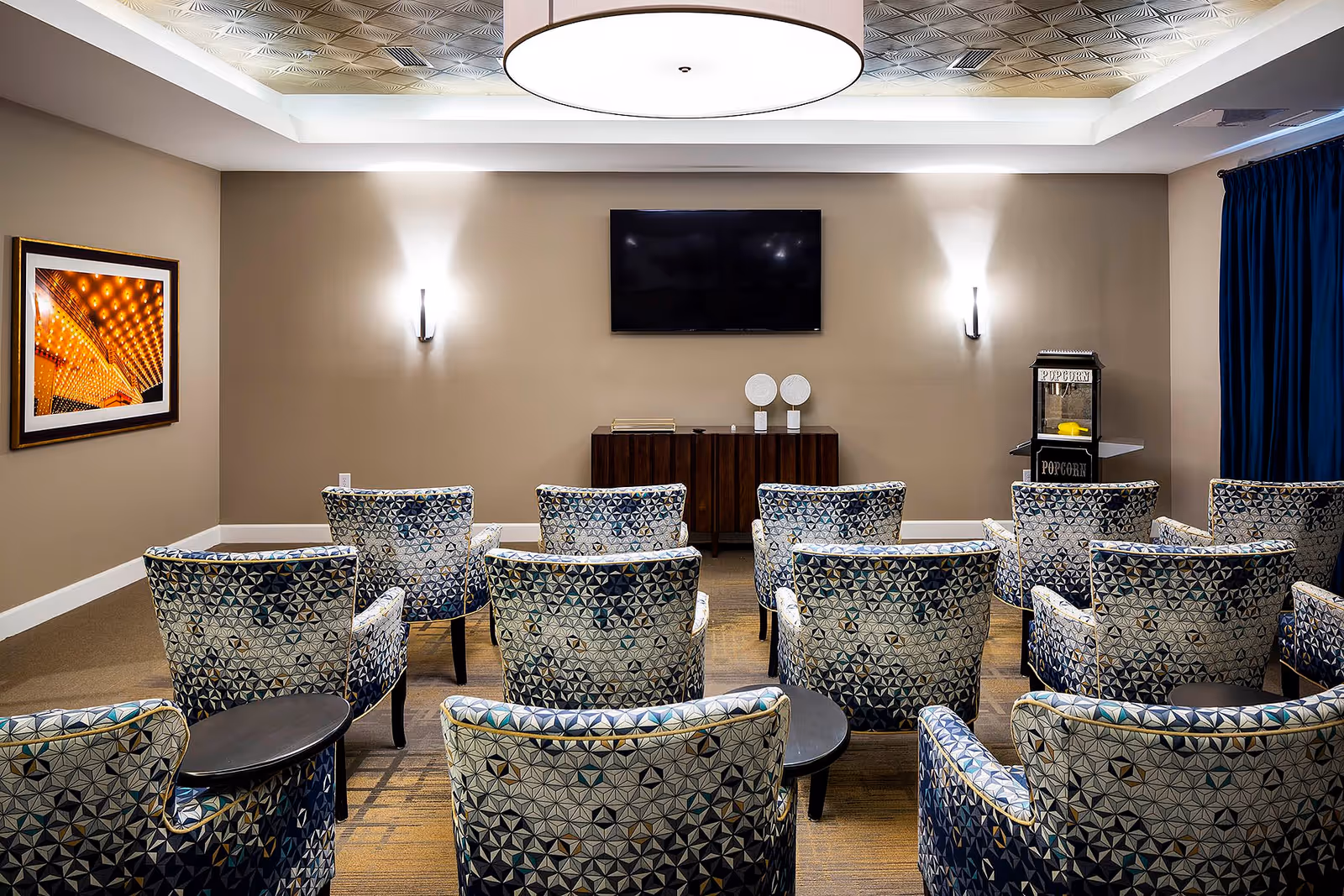 A small theater room with patterned armchairs arranged in rows facing a wall-mounted flat screen TV. The room has beige walls, a popcorn machine in the corner, a framed picture on the left wall, and a large ceiling light fixture.