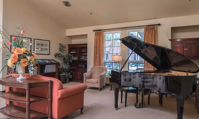 A cozy living room area in Carmel Village Retirement Community featuring a black grand piano, a red sofa, a beige armchair, a wooden side table with a floral arrangement, a window with orange curtains, and built-in wooden shelves with books and decor.