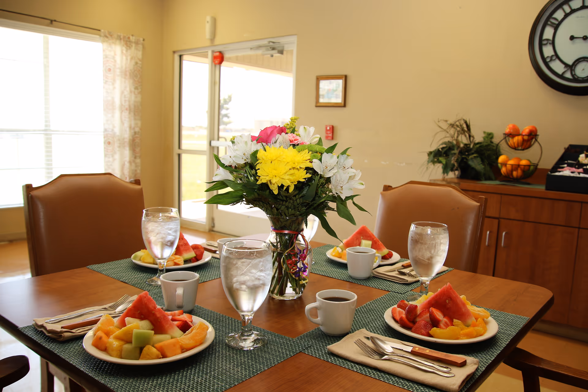 A dining table set for four with plates of fresh fruit including watermelon, strawberries, cantaloupe, and pineapple. Each place setting has a glass of water, a cup of coffee, and silverware on a green placemat. A vase with a colorful bouquet of flowers is in the center of the table. In the background, there is a wooden sideboard with a fruit basket and plants, a large wall clock, and a glass door letting in natural light.