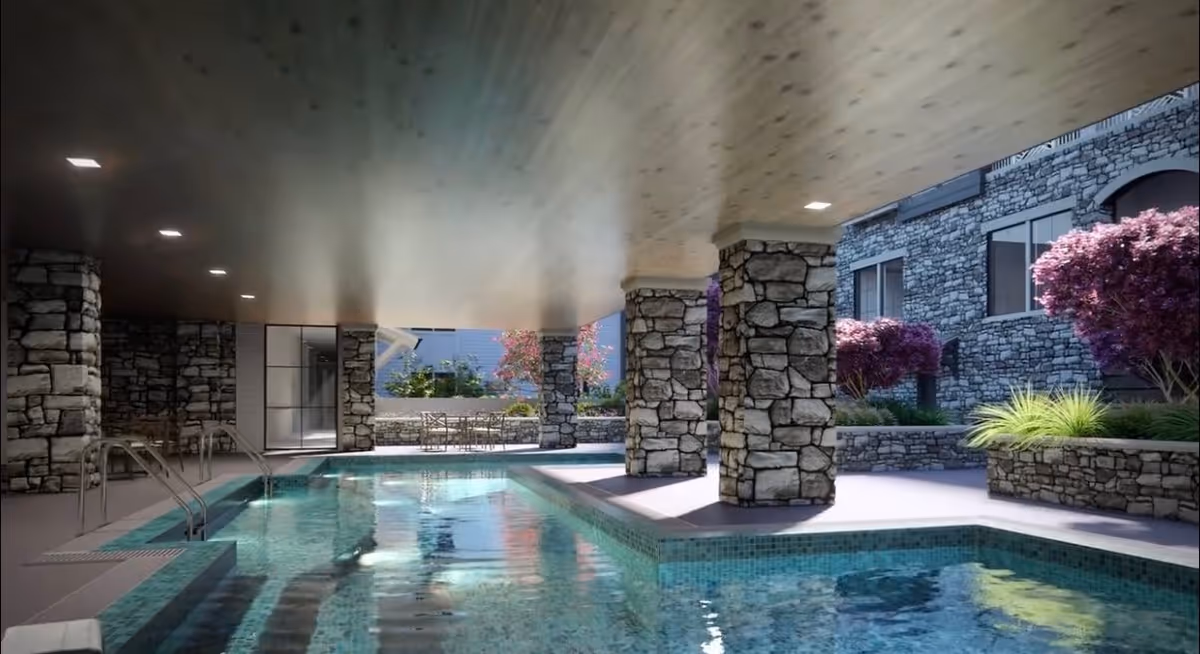 Indoor swimming pool area with stone pillars and walls, a wooden ceiling with recessed lighting, and a seating area with tables and chairs near the pool. There are plants and trees visible outside through large openings.