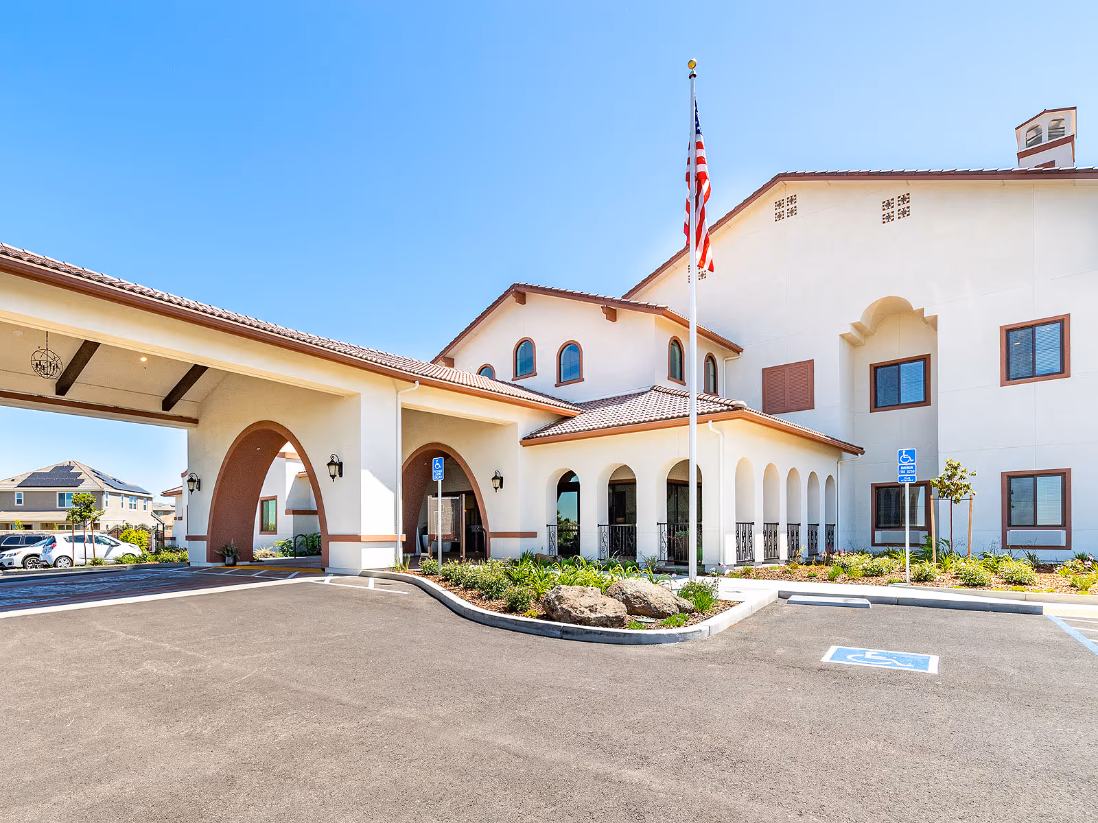 Front entrance of a Mediterranean-style senior living building with a covered porte-cochère, American flag, and accessible parking.