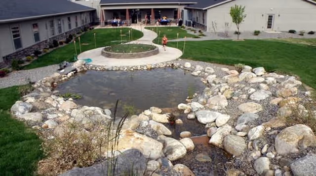 Outdoor courtyard area of a senior living facility with a pond surrounded by rocks in the foreground, a circular raised planter, green grass, and a paved walkway leading to a building with people sitting at tables under umbrellas.