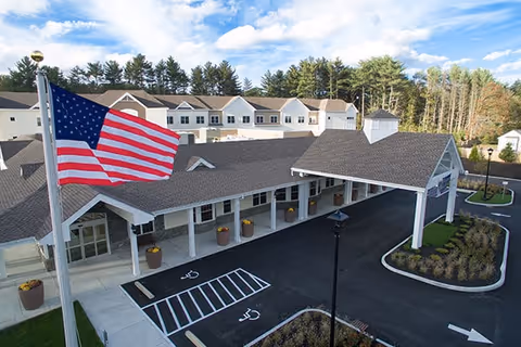 Exterior view of CareOne at Sharon senior living facility showing the main entrance with a covered drop-off area, an American flag on a flagpole, parking spaces including handicapped spots, landscaped areas, and a background of trees under a partly cloudy sky.