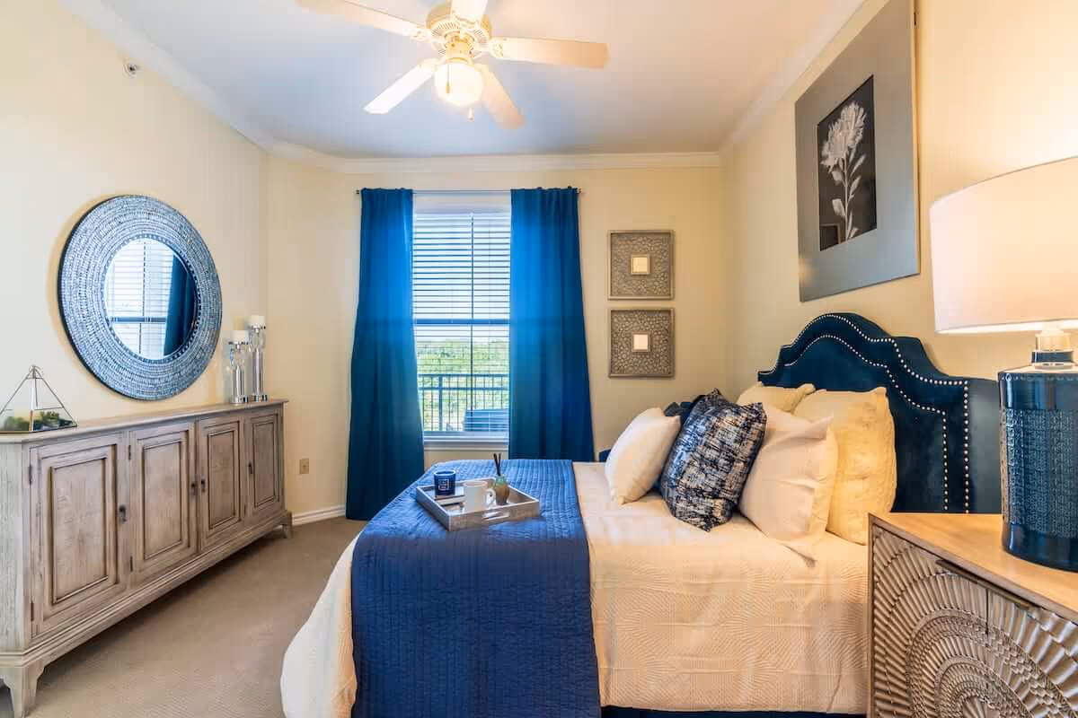 Bright staged bedroom with a blue-upholstered bed, matching blue curtains, a wooden dresser with a round mirror, and a bedside lamp.