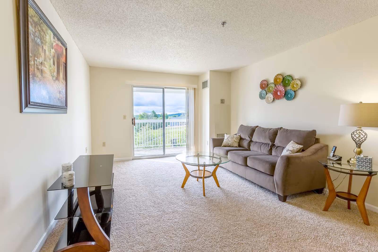A bright living room with a large gray sofa adorned with two patterned pillows, a glass coffee table with wooden legs, and a matching side table with a lamp and a phone. The room has beige carpet and walls, a colorful wall decoration above the sofa, a framed painting on the opposite wall, and a sliding glass door leading to a balcony with a view of greenery and a partly cloudy sky.