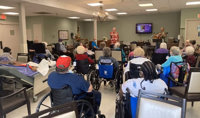Elderly residents seated in wheelchairs watch a singer and guitarist performing in a common room.