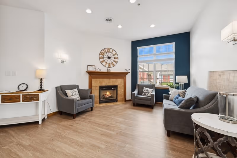 A cozy living room with a wooden floor, a fireplace with a wooden mantle, and a large round clock above it. There are two gray armchairs and a gray sofa with decorative pillows, a window with white blinds, and a small white console table with a lamp and clock on it. The walls are mostly white with one dark blue accent wall behind the window.