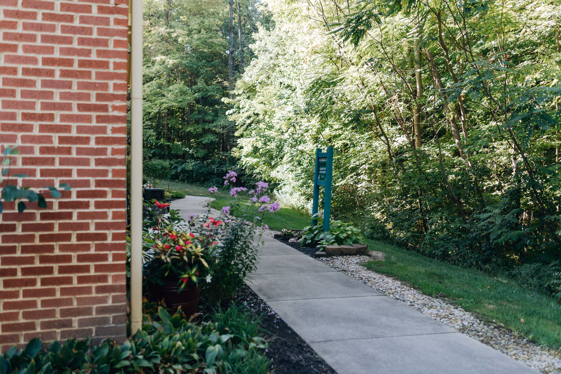 A concrete walkway beside a red brick building, bordered by green plants and colorful flowers on one side and a grassy area with trees on the other side, under bright daylight.