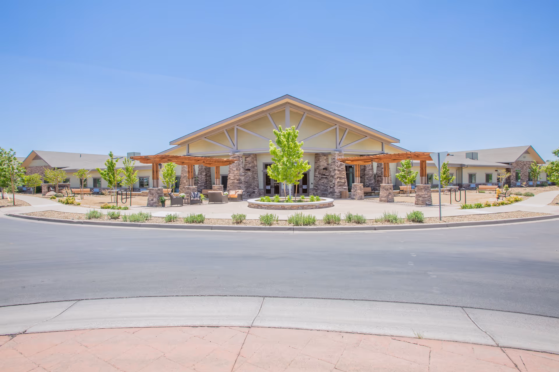 Front exterior view of Aspire Transitional Care facility featuring a single-story building with stone pillars, wooden pergolas, outdoor seating areas, young trees, and landscaped surroundings under a clear blue sky.