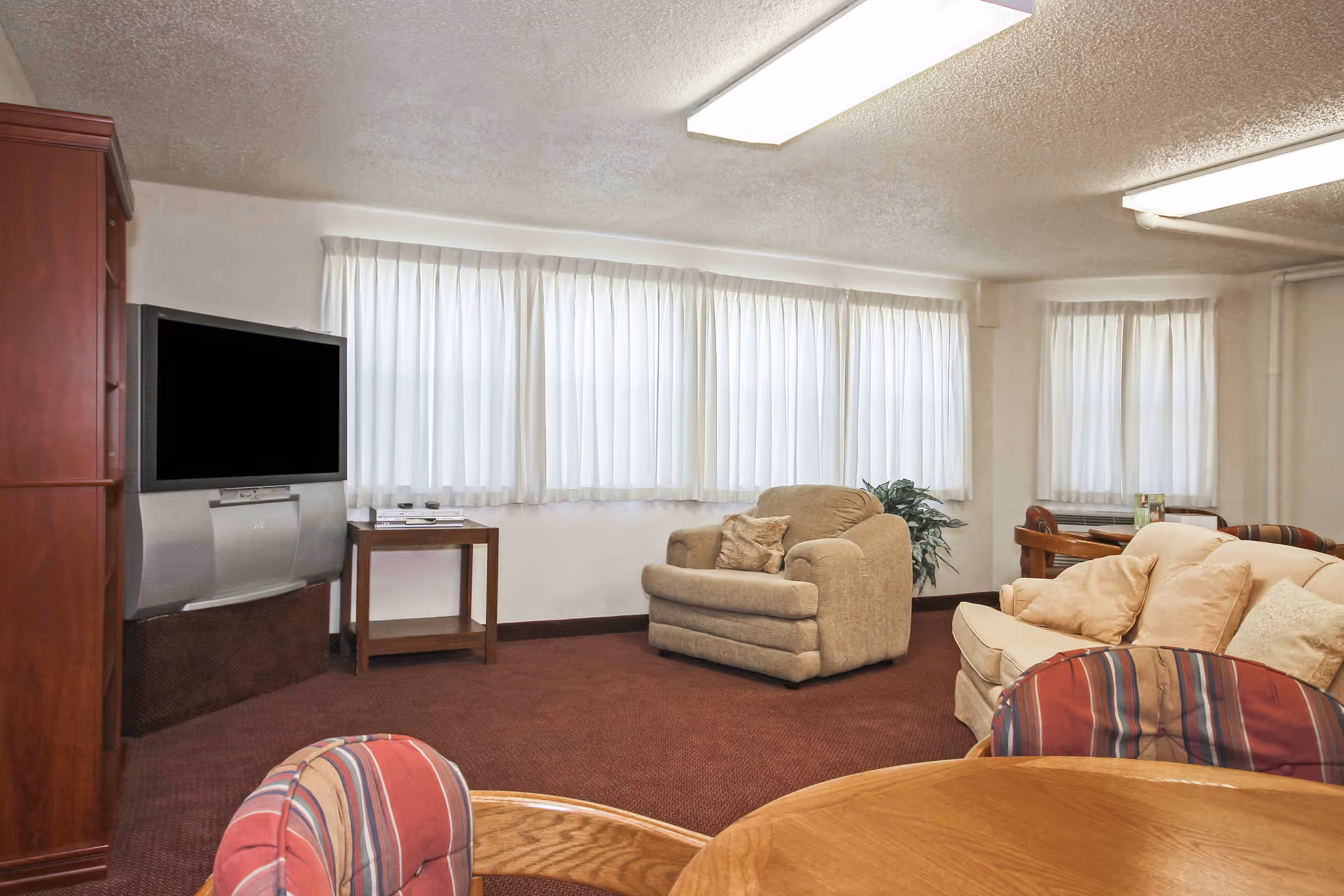 A cozy living room area with a beige armchair and sofa adorned with cushions, a wooden round table with striped cushioned chairs in the foreground, a large flat-screen TV on a stand, and windows with white curtains letting in natural light.