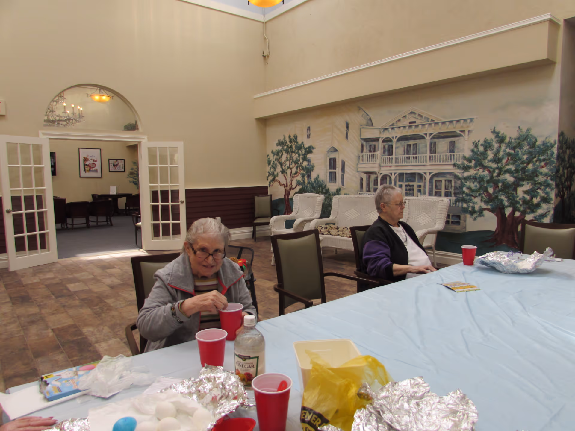 Two elderly women sitting at a large table covered with a light blue tablecloth in a spacious room. The room has a mural on the wall depicting a large house and trees. There are chairs around the table and a set of double glass doors leading to another room with framed pictures on the walls.