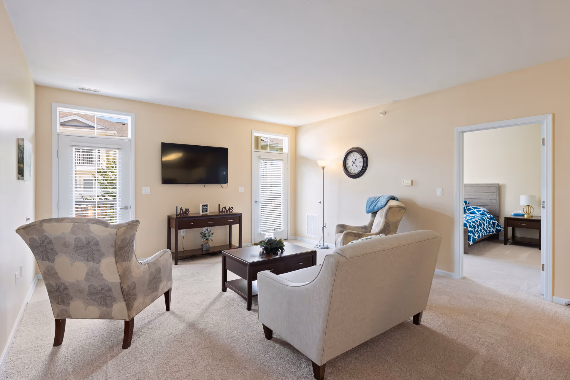 A bright living room with beige walls and carpet, featuring a beige sofa, a patterned armchair, a wooden coffee table, and a TV mounted on the wall above a console table with decorative items. There are two glass doors with blinds letting in natural light, a floor lamp, and a wall clock. A doorway leads to a bedroom with a bed and nightstand visible.
