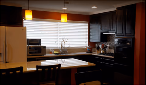 Interior view of a kitchen with dark wooden cabinets, a refrigerator, microwave, oven, and a countertop island with chairs. Two pendant lights hang from the ceiling above the island, and a window with closed blinds is above the sink.