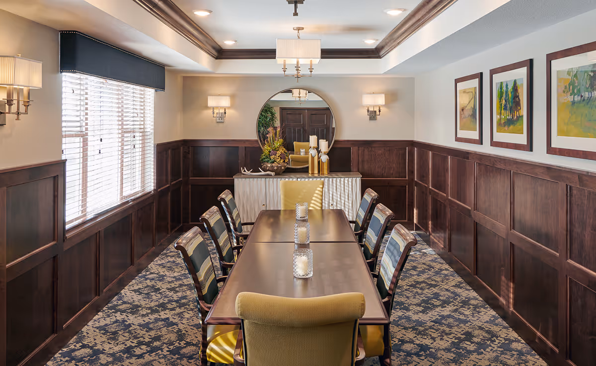 Long dining room with a polished wooden table and upholstered chairs, wood-paneled walls, a chandelier, and framed artwork.