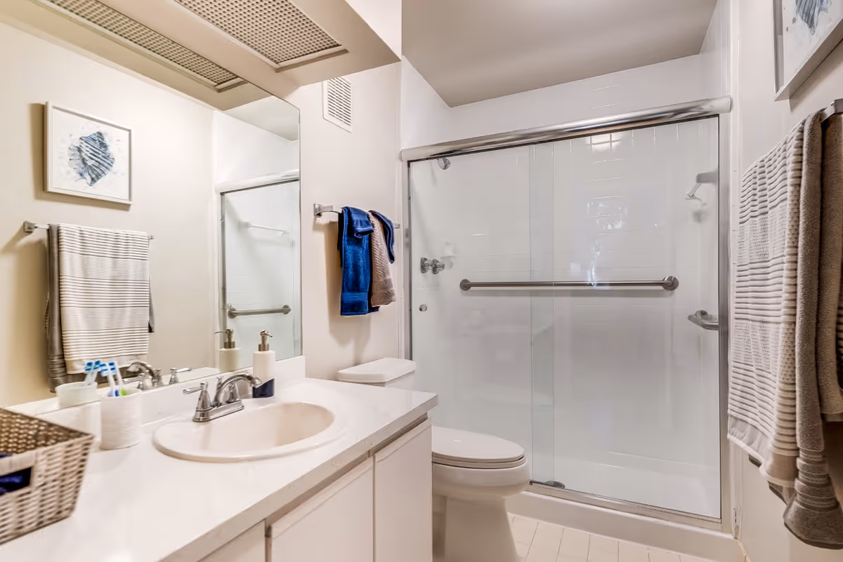 A clean and modern bathroom featuring a white countertop with a sink, a large mirror above the sink, a toilet, and a glass-enclosed shower with a metal grab bar. There are striped towels hanging on the wall and a small framed artwork above the towel rack.
