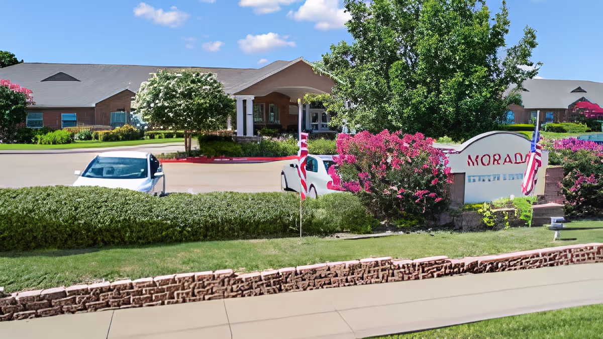 Exterior view of Morada Cedar Hill facility showing a brick building with a covered entrance, surrounded by green trees, bushes, and flowering plants. Two cars are parked near the entrance, and an American flag is displayed on a pole near a sign with the facility's name.